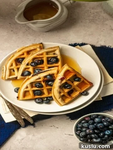 Blueberry waffle on a plate on top of a book and blue napkin with silver forks and a syrup pourer.