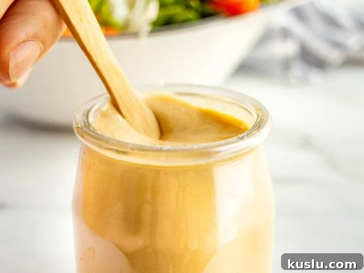 A glass jar with thick light brown dressing in it being stirred by a hand