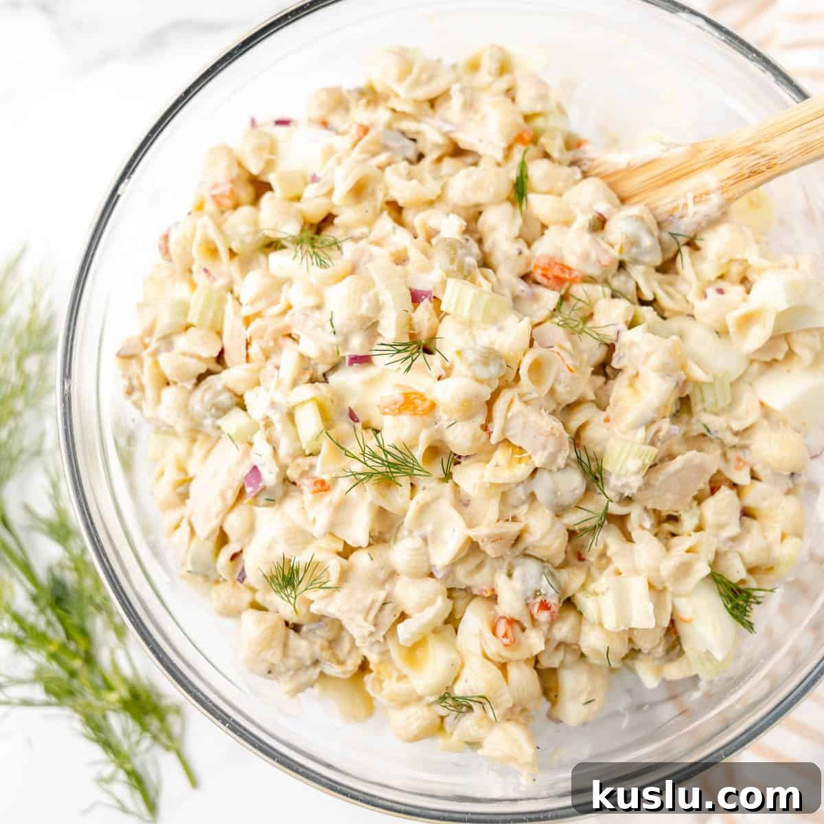 Mediterranean Tuna Pasta Salad with Olives and Cherry Tomatoes 10 A glass bowl filled with creamy pasta salad containing shell pasta, diced vegetables, and fresh dill, with a wooden spoon resting inside the bowl.