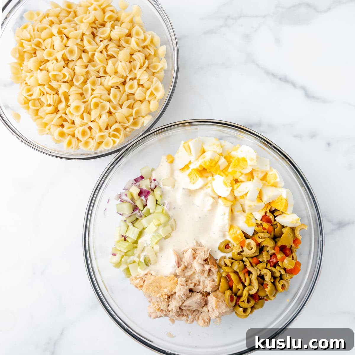 Mediterranean Tuna Pasta Salad with Olives and Cherry Tomatoes 3 Two glass bowls on a marble surface; one contains cooked shell pasta, the other contains chopped celery, hard-boiled eggs, olives, tuna, red onion, and a creamy dressing.