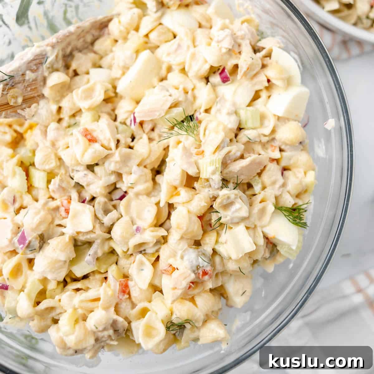 Mediterranean Tuna Pasta Salad with Olives and Cherry Tomatoes 4 A glass bowl filled with creamy pasta salad containing shell pasta, chopped vegetables, herbs, and diced hard-boiled eggs, with a wooden spoon in the mixture.