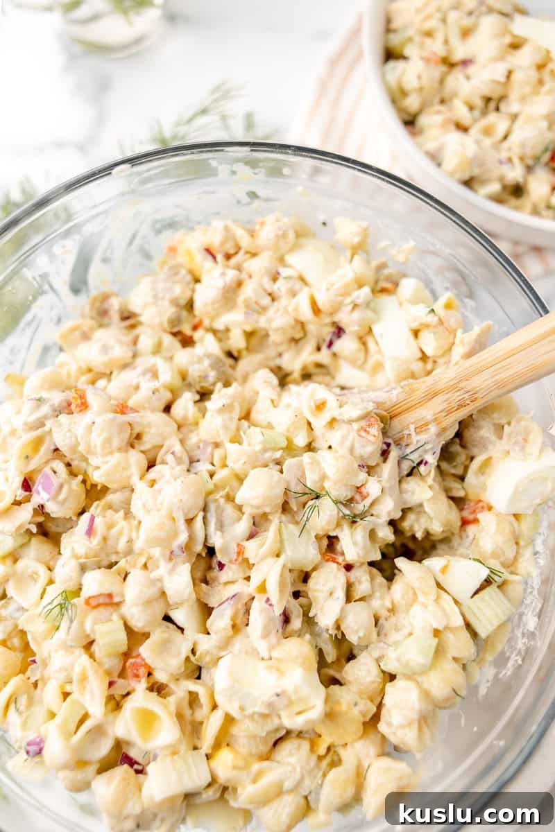 Mediterranean Tuna Pasta Salad with Olives and Cherry Tomatoes 5 A glass bowl filled with creamy pasta salad containing shell pasta, chopped vegetables, and herbs, with a wooden spoon for mixing.