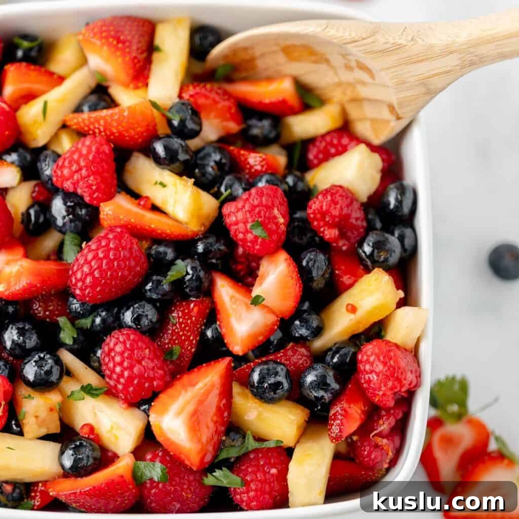 Mediterranean Tuna Pasta Salad with Olives and Cherry Tomatoes 8 A white bowl filled with a fruit salad containing strawberries, raspberries, blueberries, pineapple chunks, and garnished with herbs. A wooden spoon rests on the side.