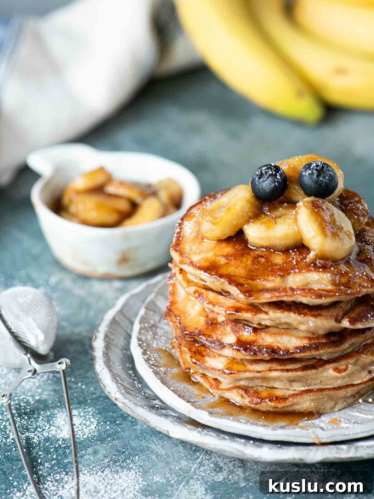 A stack of pancakes with bananas on top placed on grey plate over a blue surface.