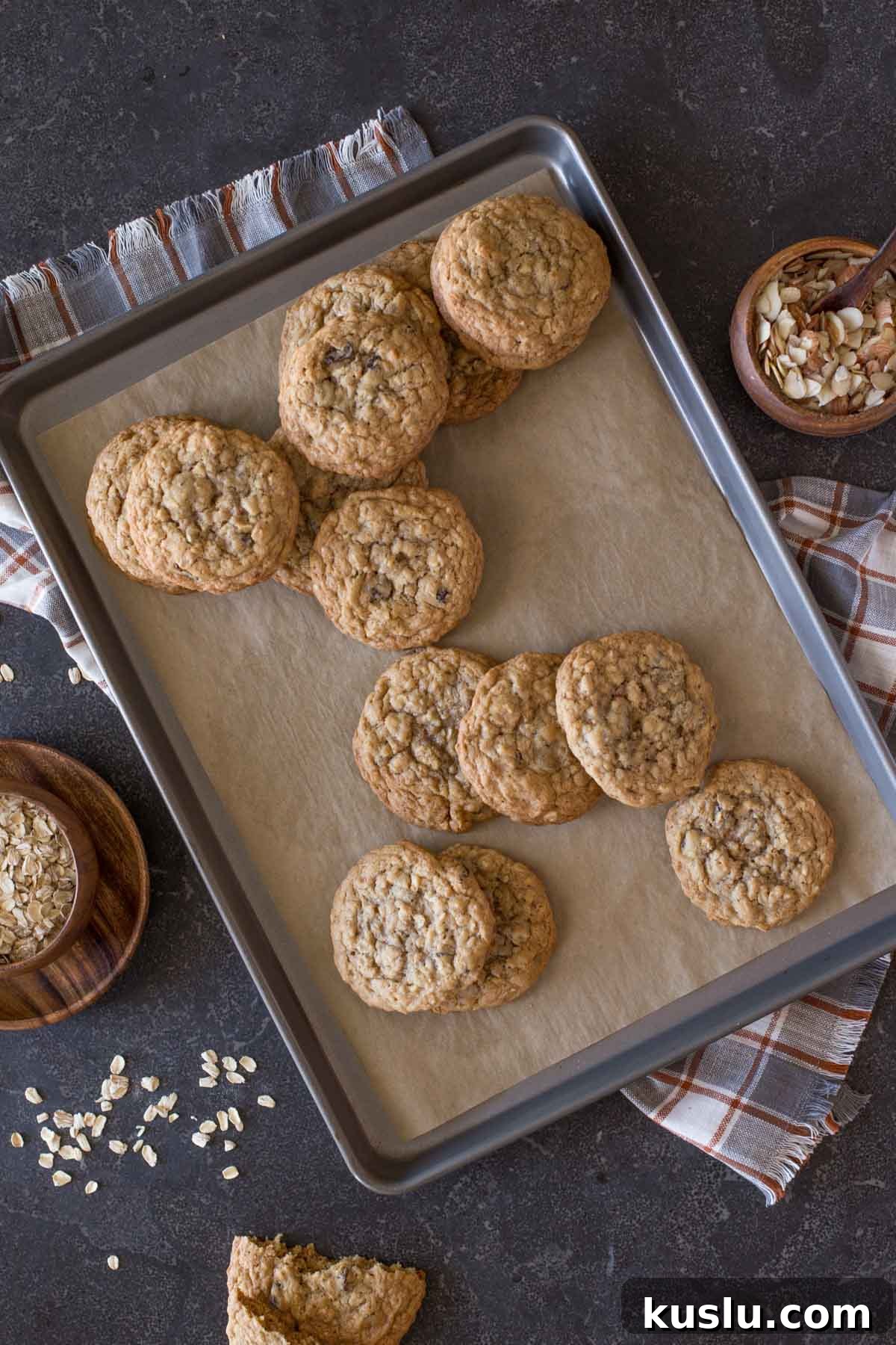 Bakery Style Oatmeal Raisin Cookies on a parchment paper lined baking sheet, with a small wood bowl of oats on one side of the baking sheet, and a small wood bowl of slice almonds on the other side. 