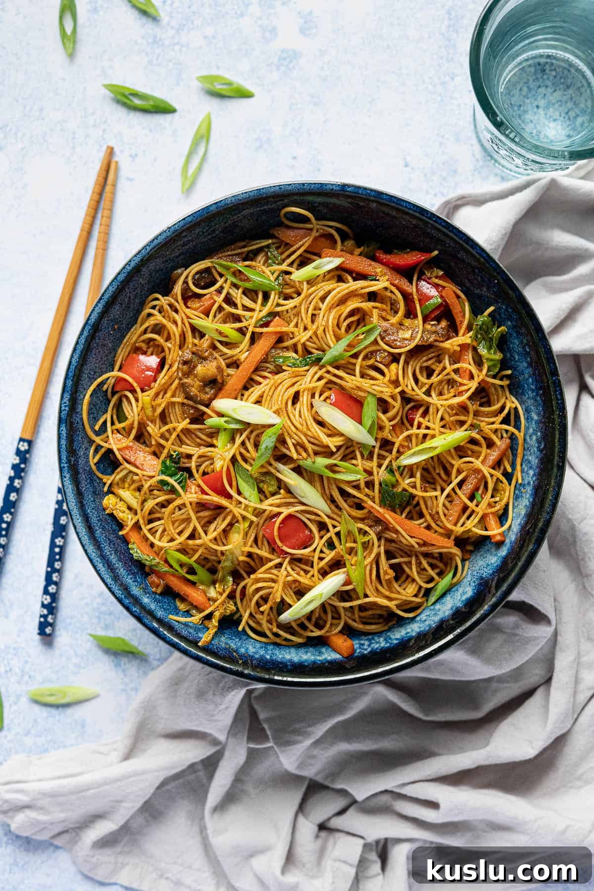 A blue bowl of vegan curry noodles, surrounded by chopsticks, a grey cloth, glass of water and sliced spring onions.