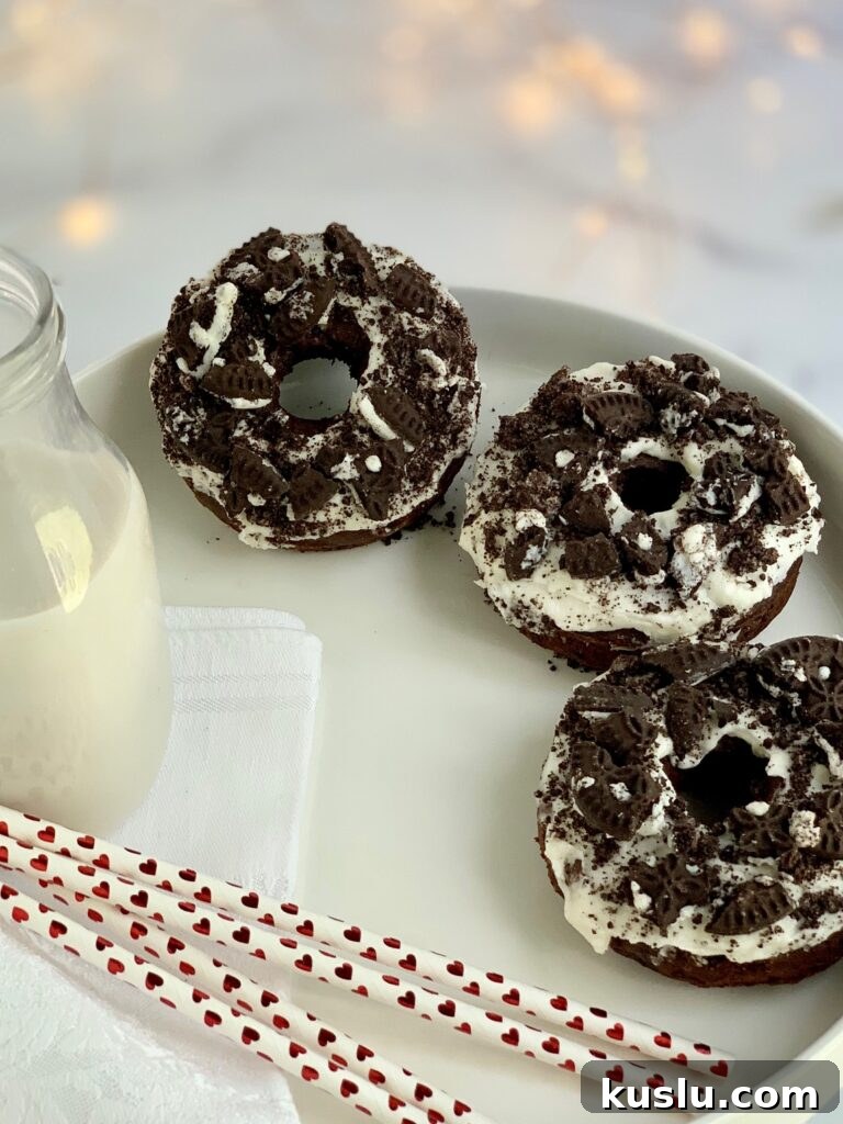 3 oreo donuts with a creamy white frosting and oreo crumbs sprinkled on top next to a glass of milk and 3 straws with red hearts on them.