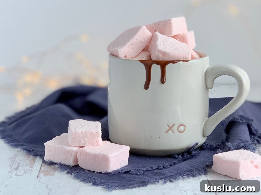Soft fluffy pale pink square marshmallows on a table next to a mug of hot chocolate. 