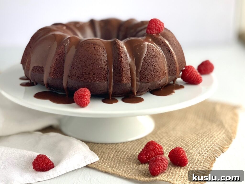 A large chocolate bundt cake on a white serving cake plate with a chocolate ganache running down the sides and random strawberries throughout. 