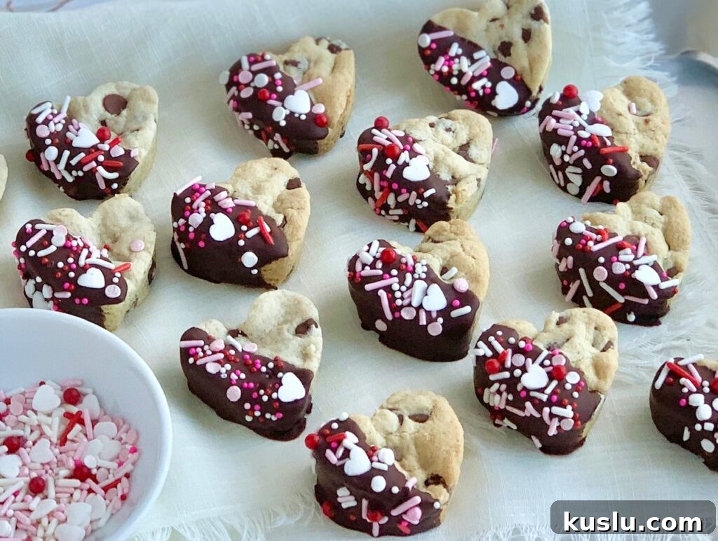 4 rows of mini heart shaped chocolate chip cookies that have been dipped halfway into melted chocolate then topped with pink, white, and red sprinkles.